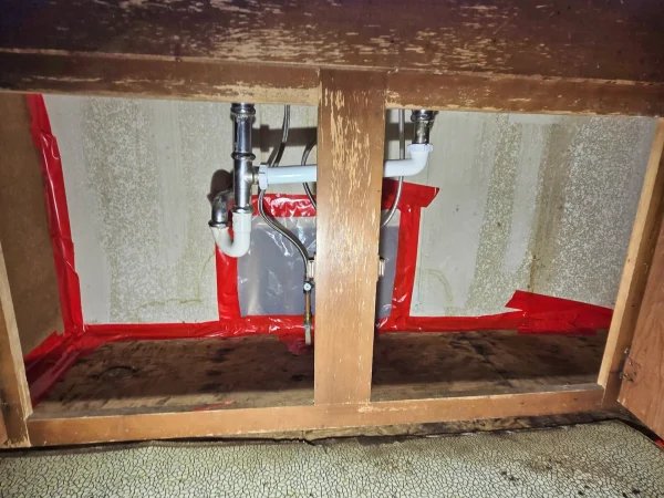 Exposed wall and subfloor inside a kitchen cabinet during the plumbing rough-in phase.